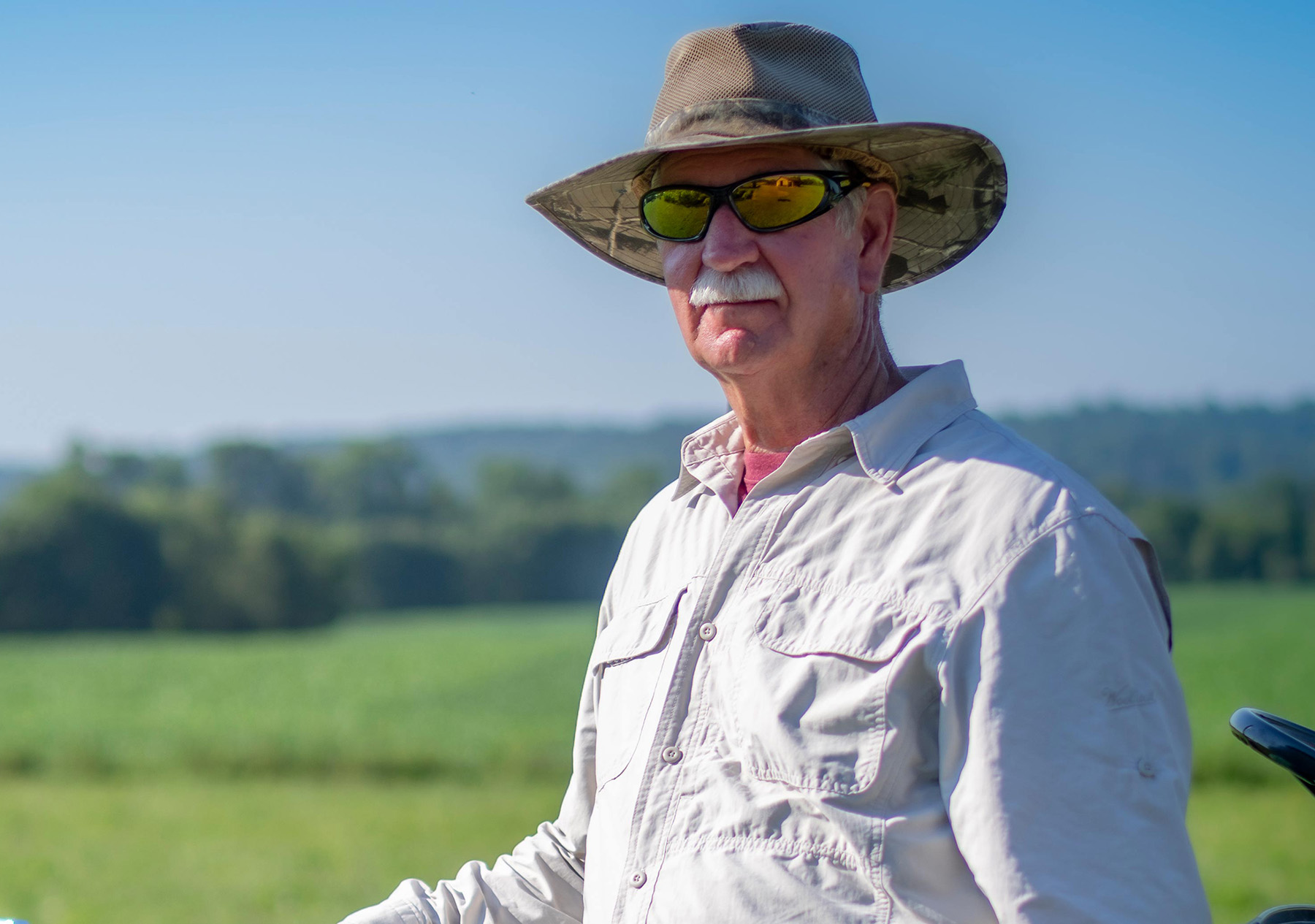 Carl H. stands in a sunny field wearing a hat and sunglasses.